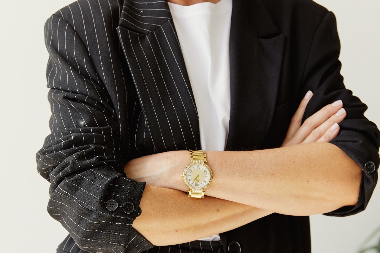 a woman in a blazer with her arms crossed, wearing a diamond studded watch