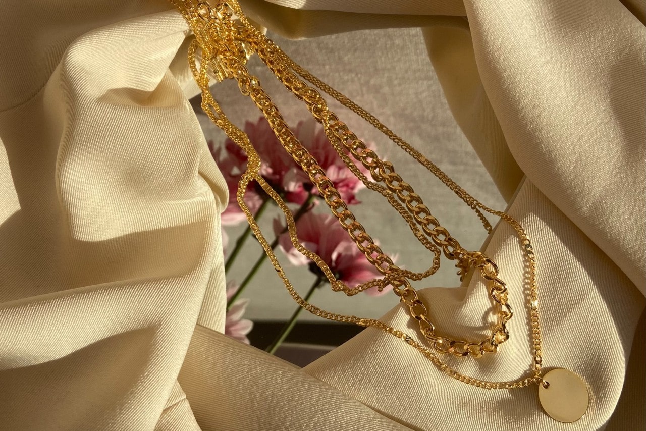 A trio of gold necklaces sitting on a mirror surrounded by tan fabric.