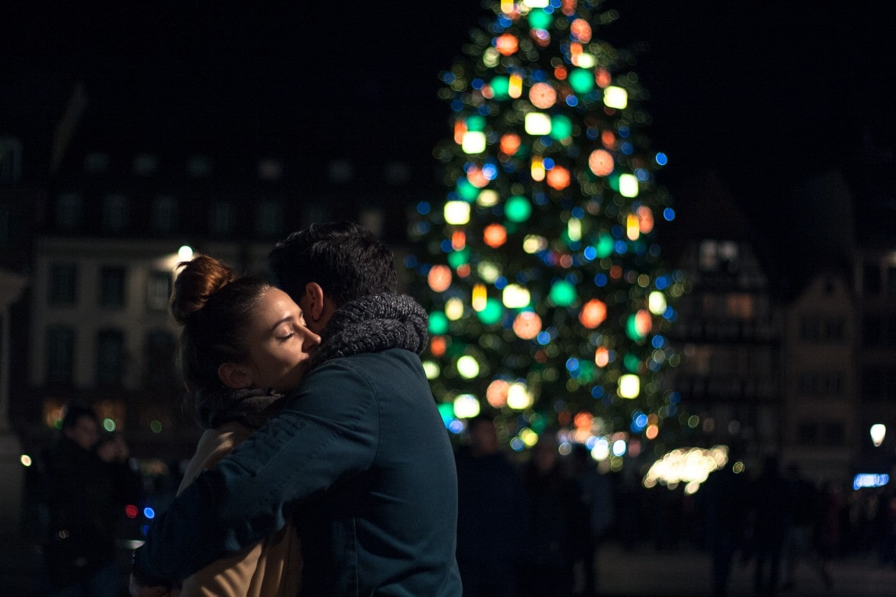 A couple embraces in front of a lit Christmas tree outside.