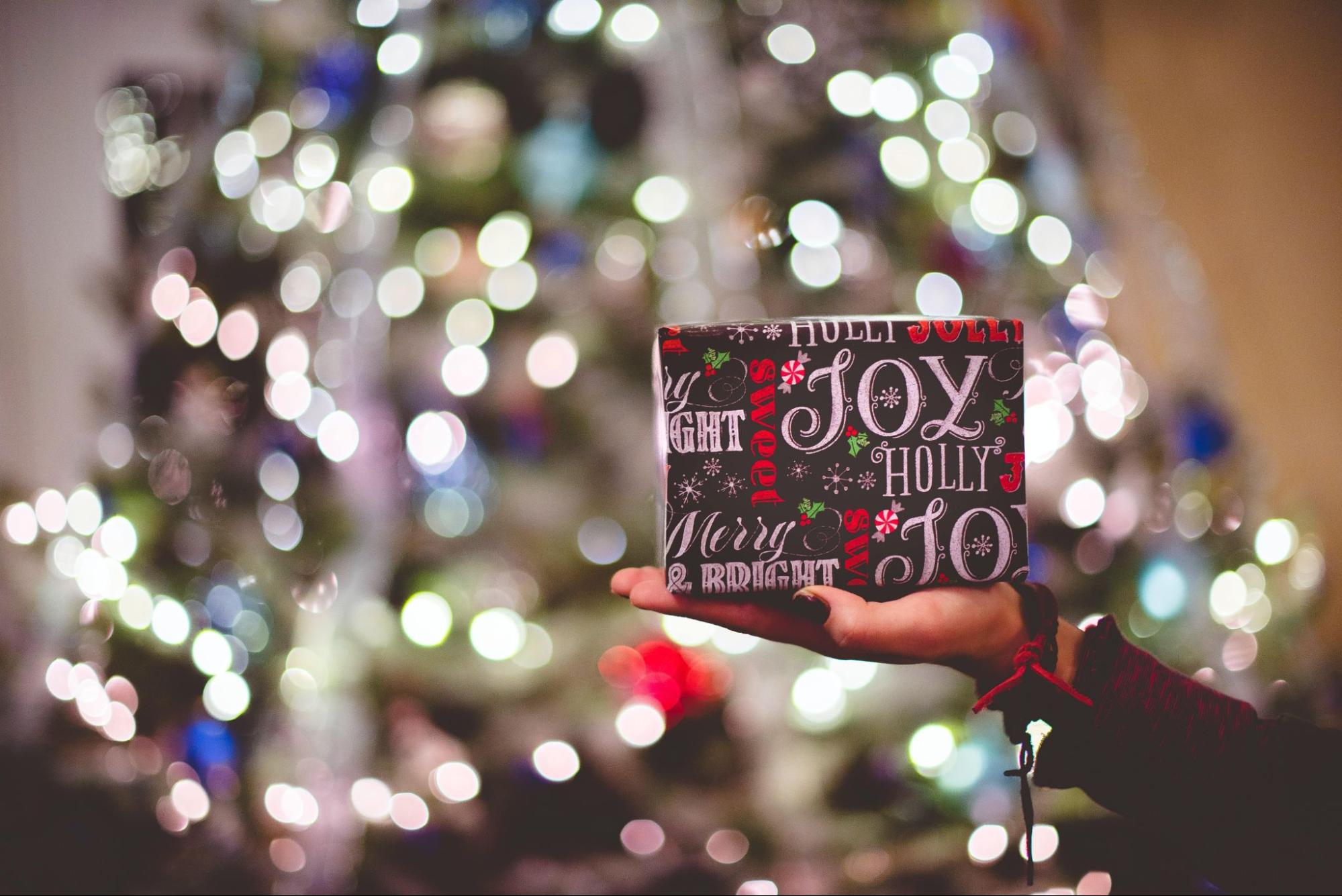 A woman holds a wrapped gift in her hand in front of a lit Christmas tree.