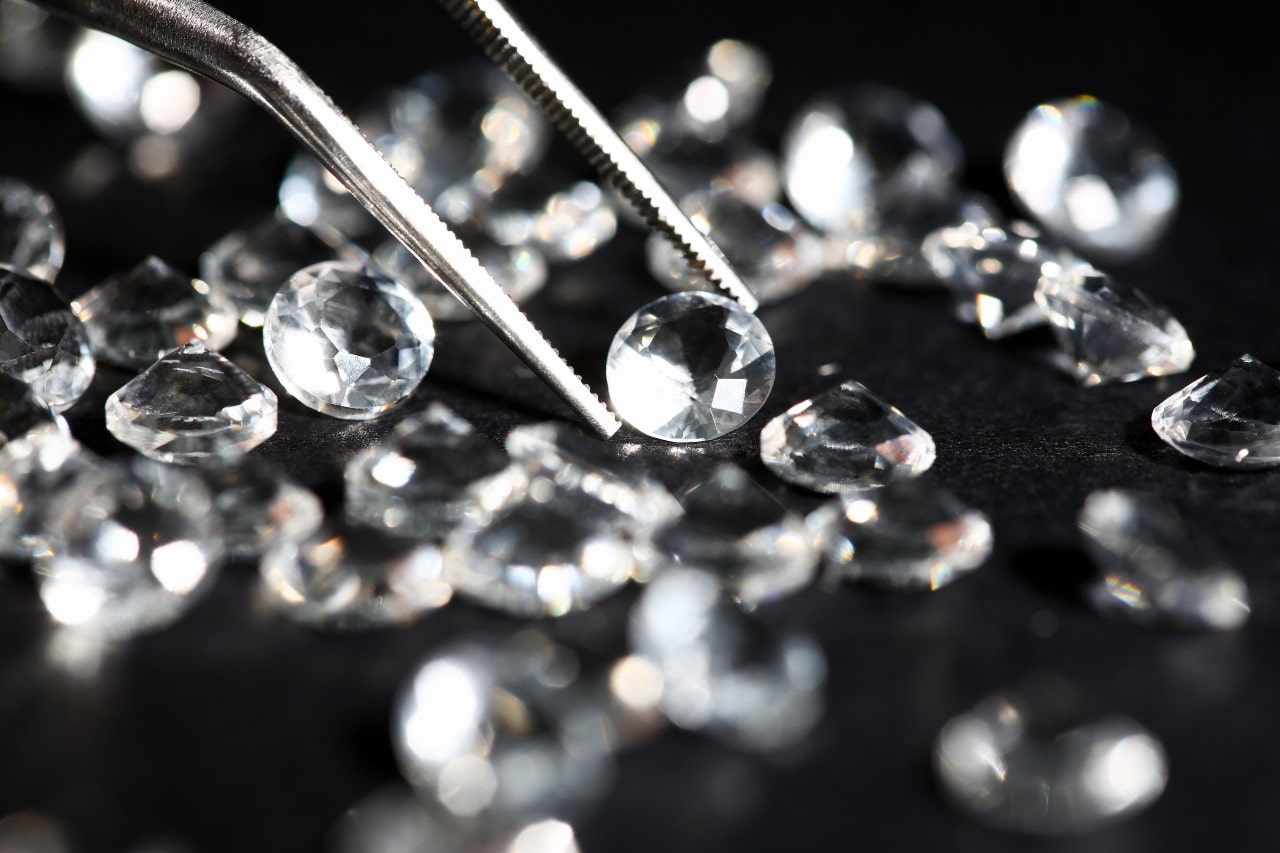 A pair of jewelry tweezers selects a diamond from an array of round-cut diamonds.