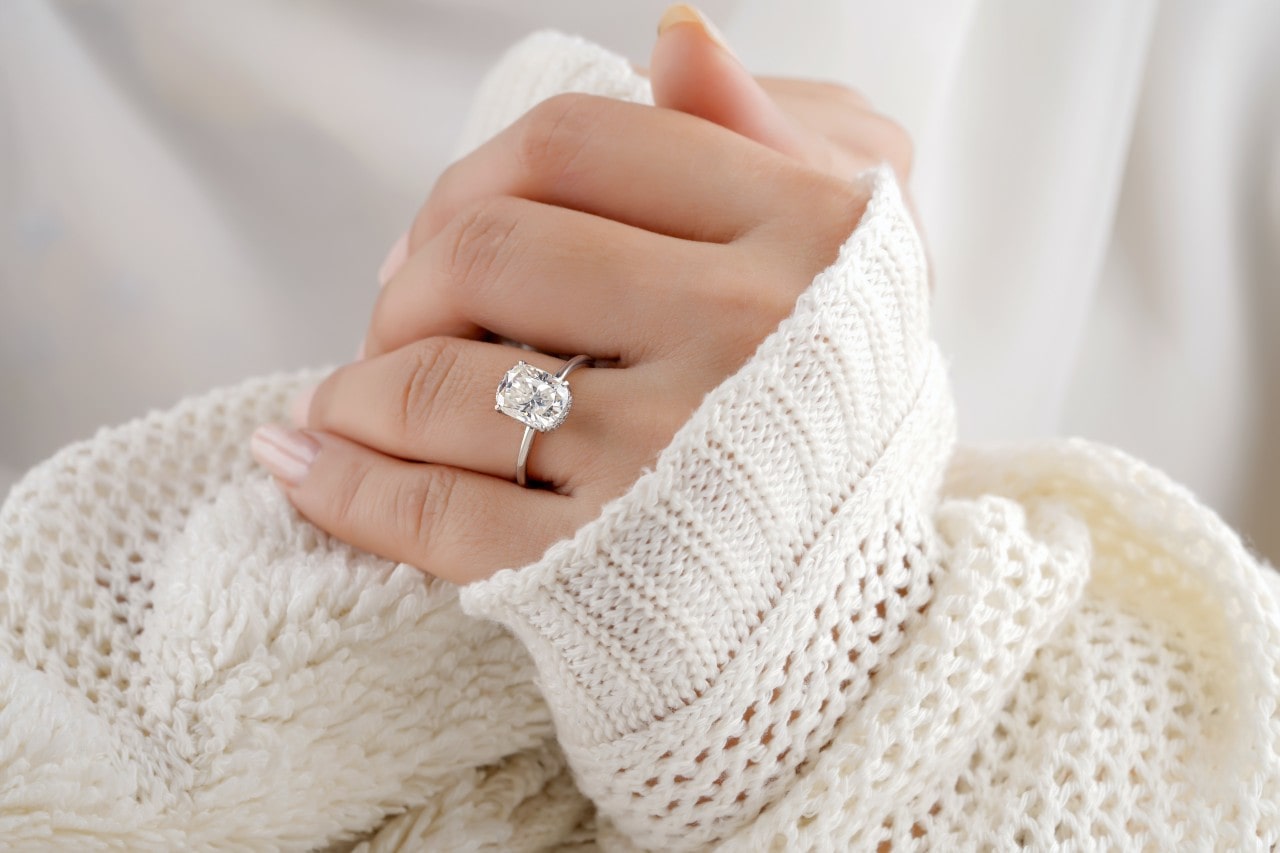 A woman’s hands wearing a chunky white sweater and an oval-cut engagement ring